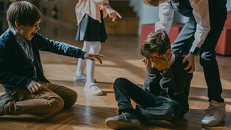 EinJunge sitzt auf dem Boden und versteckt sein Gesicht mit den Händen. Zwei Kinder zeigen mit dem Finger auf ihn. Ein drittes Kind steht hinter ihm und fasst ihn an der Schulter.