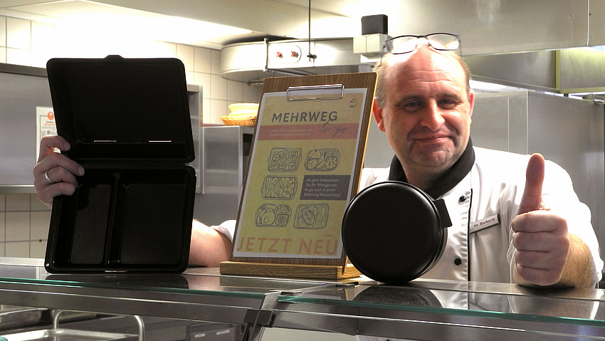 Ein Mann steht hinter dem Tresen einer Kantine. In der rechten Hand hält er eine Mehrwegverpackung, mit der linken Hand zeigt er mit dem Daumen nach oben.