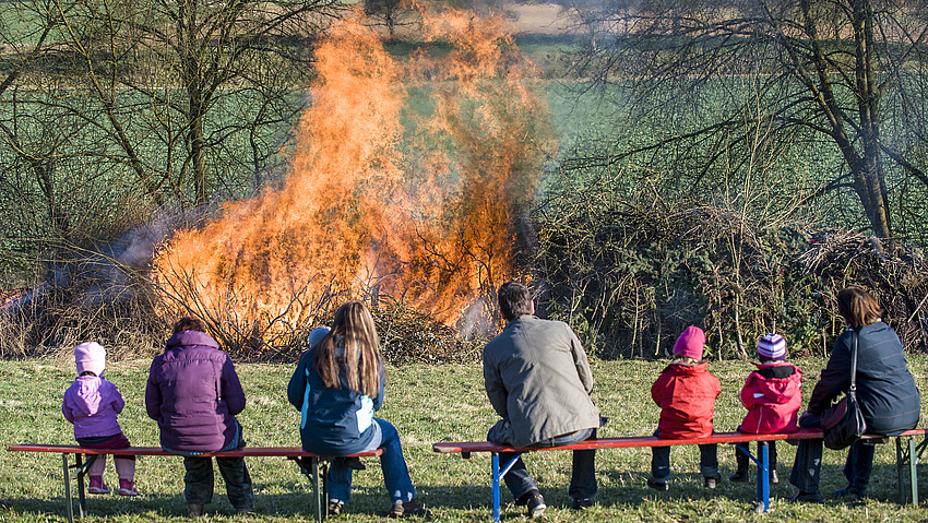 Mehrere Personen sitzen auf Bänken und sehen einem Feuer zu.