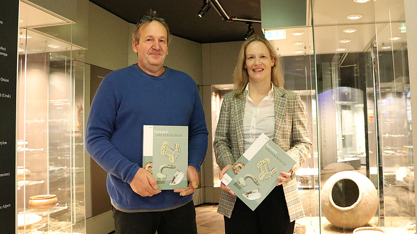 Zwei Menschen stehen vor Vitrinen in einem Museum und halten jeweils zwei Bücher in der Hand.