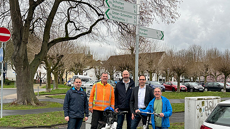 Fünf Männer mit Fahrrädern stehen auf einer Straße. Im Hintergrund sind Radwegebeschilderungen zu sehen.