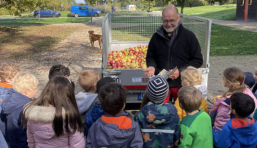 Uli Schwabe vor einem Wagen mit Äpfeln. ER hält einen Apfel in der Hand und spricht mit einer Gruppe von Kindern