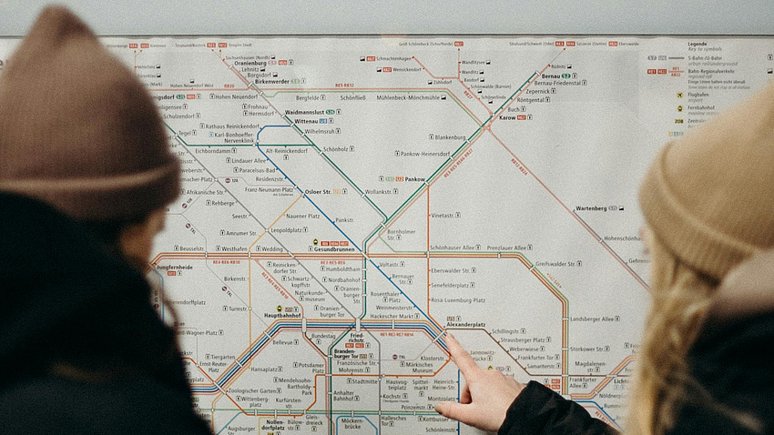 Zwei Personen stehen vor einem U-Bahn Fahrplan und die Person rechts im Bild zeigt auf einen Haltepunkt.