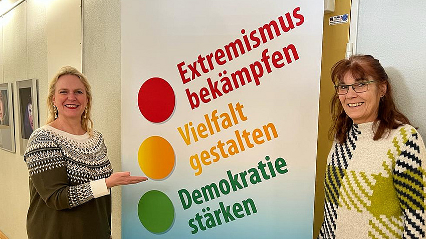 Zwei Frauen stehen vor einem Roll-up mit der Aufschrift "Extremismus bekämpfen, Vielfalt gestalten, Demokratie stärken"