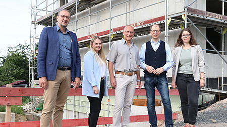 Fünf Personen stehen auf einer Baustelle vor dem Rohbau eines Hauses.