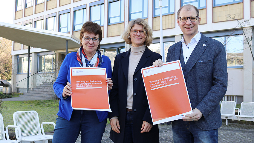 Zwei Frauen und ein Mann stehen nebeneinander und lächeln in die Kamera. Zwei von ihnen haben ein kleines Plakat in der Hand. Auf diesem steht: Verhütung und Bekämpfung von Gewalt gegen Frauen und häuslicher Gewalt.